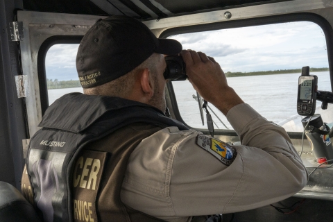 A Federal Wildlife Officer uses binoculars at the helm of a boat with the shoreline visible through the window. 