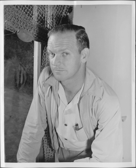 Portrait of a man, taken from the waist up; he wears a button-up shirt, and a light-weight jacket. To the left of him is what appears to be a window with a mesh curtain or drape.