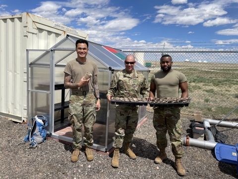 Three military members hold trays of milkweed plugs outside