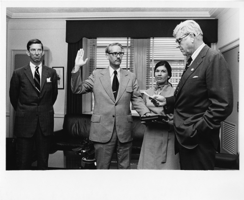 A scene of four people in an office-setting; the man in the center is taking an oath, and has his right hand raised in the air, and his left hand resting on a book that is being held by a woman standing to the right of him. The man on the right is turned to the side, and is reading from a card or small piece of paper he's holding in his right hand. Another man standing on the left, with his hands behind his back. The three me are wearing business suits and ties, while the woman wears a light dress