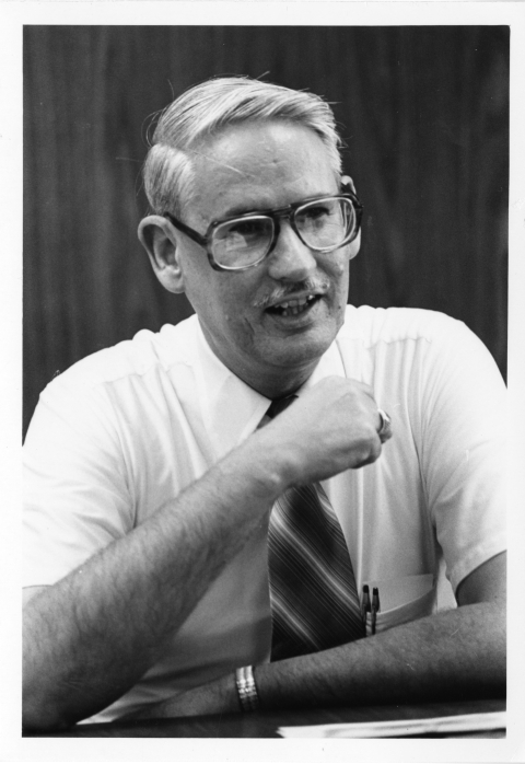 Close-up of a man sitting at a desk or table, with his right arm leaning on the table, and his left arm held across his chest. He wears a light-colored collared dress shirt with short sleeves, and a necktie with diagonal stripes. The tips of two pens are visible in his shirt pocket. He also wears horn rimmed eyeglass, and appears to be in the middle of saying something