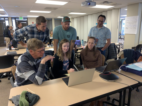 Six participants in a classroom all look at one person's laptop
