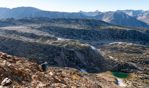 A biologist walking downhill on a steep slope with large mountains in the background.