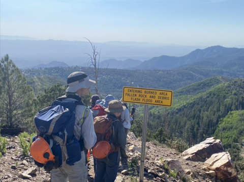 A crew hikes in a field by mountains, next to a sign that reads "Entering Burned Area Fallen Rock and Debris Flash Flood Area"