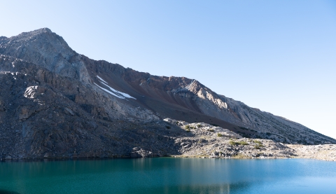 Bright blue lake in the foreground. A steep, rocky mountain sits behind the lake