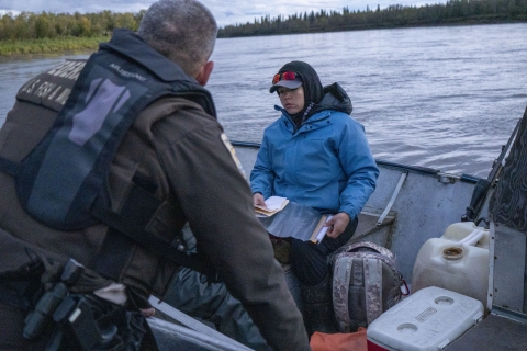 A uniformed officer on a boat holds on to another boat with a moose hunter as she gets her license ready as both boats float in the middle of a large river with a wooded shoreline in the background.