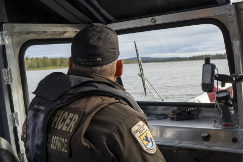 uniformed officer looks out of the covered helm areas of a river boat with the shoreline visible through the windshield. 