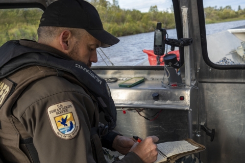 A uniformed officer takes notes during a patrol on a boat with the shoreline visible outside the windshield of the cabin. 