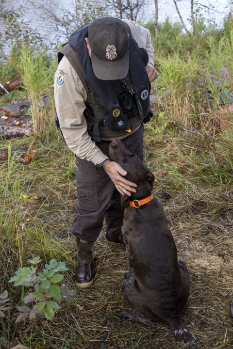 A uniformed officer near the bank of a river looks down and pets a dog that is seated in front of him. 