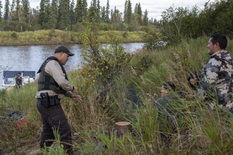 A uniformed officer at the edge of thick vegetation talking to a family of hunters sitting in the brushy area with a large river in the background and a boat on the riverbank with another officer near the helm on a satellite phone. 