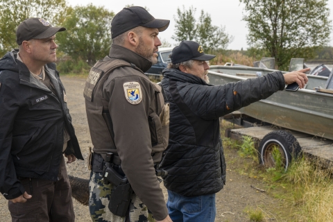 two uniformed officers stand with a person as the person points at something and the officers looking in the same direction. The group stands in front of a boat on a trailer. 