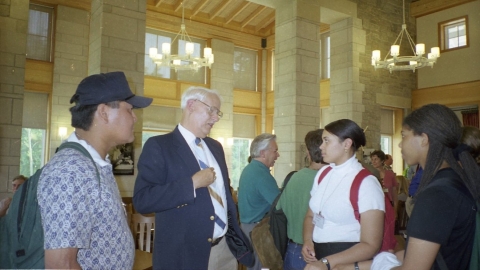Older man, center, having a conversation with three younger people (one man, left, and two women, right), in a large room with a high ceiling, and several chandeliers, as well as large, floor-to-ceiling windows. The older man is dressed in a blazer, dress shirt, and necktie, while the younger people are more casually dressed (e.g. the man on the left wears a baseball cap); they also have backpacks on.
