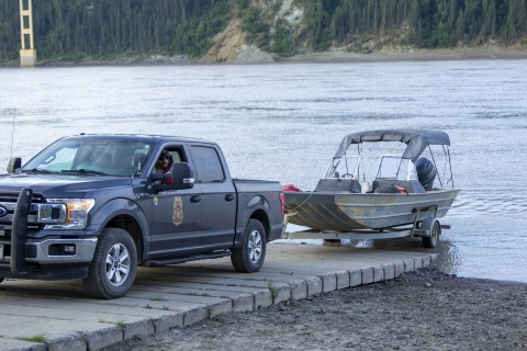 A Federal Wildlife Officer in a Refuge Law Enforcement truck backs a trailer with a boat down a boat launch ramp into a large river with a bridge in the background