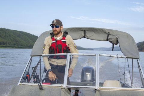 A Federal Wildlife Officer stands behind the helm of a boat in the middle of a large rive river. 