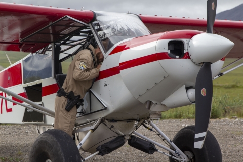A uniformed officer stands under the wing of a top cub bush plane reaching into the cockpit and a flat tundra landscape in the background with mountains of the brooks range of Alaska in the horizon. 