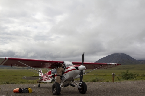A uniformed officer stands under the wing of a top cub bush plane reaching into the cockpit with three small bags on the floor nearby and a flat tundra landscape in the background with mountains of the brooks range of Alaska in the horizon. 