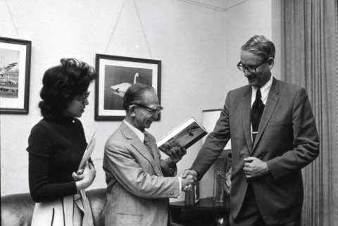 Three people posing together - man in the center holds up a hardcover book in his left hand, while shaking the hand of a taller man on the right; both wear business suits and eyeglasses. A woman stands on the left, holding a batch of papers against her chest. She's dressed in a light-colored skirt, and a dark-colored top. She also wears eyeglasses. Visible behind them are a couch, and an end table that features a couple of books and a table lamp. Framed artworks hang on the wall behind them