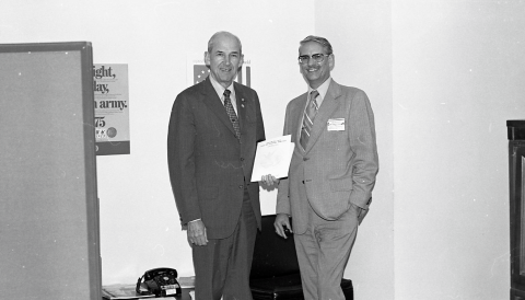Two men in business suits, posing together for a photograph. The man on the left (Joh Gottschalk) is holding an award certificate of some kind in his left hand. The man on the left is balding, and wears a dark-colored suit, while the man on the right has a mustache and wears a light-colored suit, as well as a pair of eyeglasses. Directly behind them is a low desk or table, with a telephone of 1970s vintage on the tabletop. Two posters appear on the wall behind them.