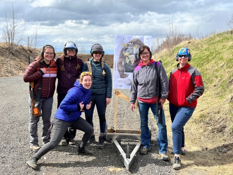 six people pose next to a bear target on a cart with wheels. A couple of them carry shotguns slung over their shoulders. Shooting range berms in the background.
