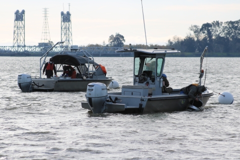 Two boats float on a river while a man leans out of one boat and dunks a large, cylindrical object in the water