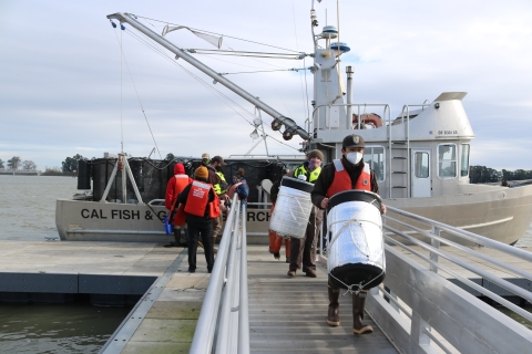 Two people carry large cylindrical objects away from a boat on a dock.