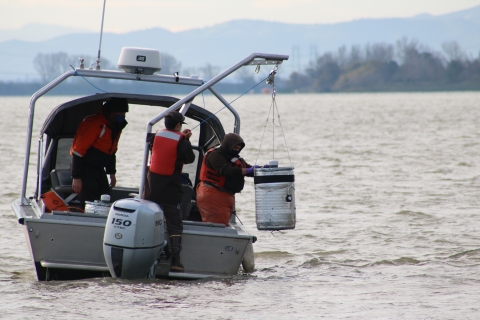 A woman on a boat lowers a large, cylindrical object into a river with assistance from a crane-like device.