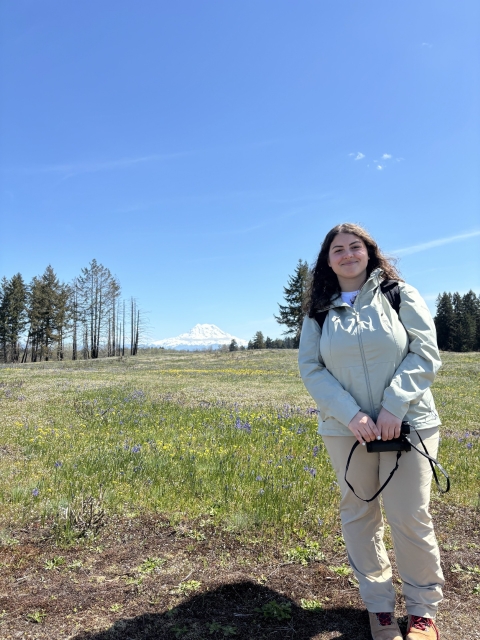 A young woman smiles and holds a pair of binoculars. She stands in a grassland full of wildlfowers, and a large snow covered mountain is visible on the horizon. 