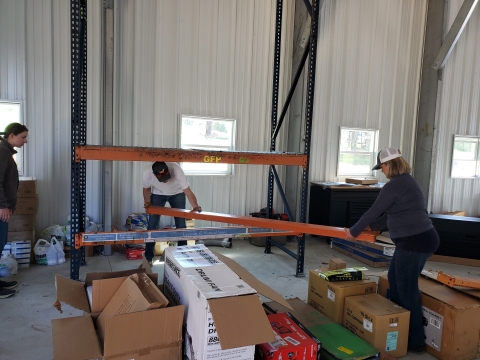 Three people building industrial shelving. Background is evenly spaced windows at chest height. Foreground has a variety of partially opened boxes of varying size