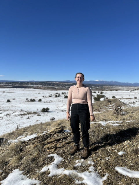 A young woman stands and smiles in a snowy plain, with a clear sunny sky and a large mountain range visible in the background. 