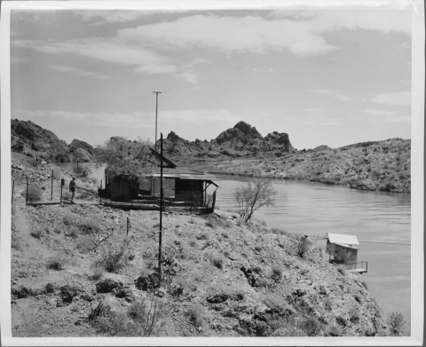 View from the bank looking over into a large lake; in the center on top of the bank is a rough hewn wooden structure (watchman's cabin), with a crudely constructed small building just below it, right along the shore. A man stands just to the left of the center structure. A few low mountain peaks are on the horizon. The environment is of an arid desert, with scrubby vegetation scattered throughout.