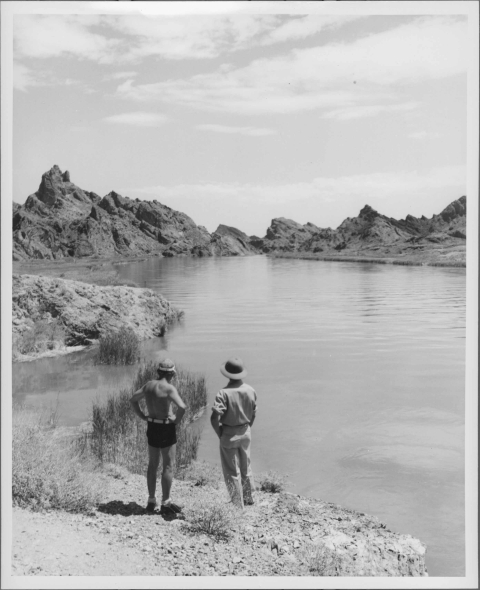 Two men in the foreground, standing with their backs to the camera, looking out over a large lake. The man on the left is shirtless, but wears a cap; the man on the right appears to be be wearing a uniform shirt and pants, as well as a sun helmet. A mountain ridge appears on the horizon, at the opposite end of the lake.