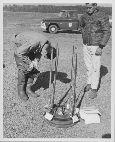 Two men standing around a device made to hold leg bands for banding birds, made from the metal hub of a tire and about 6 tall metal rods that hold the metal bands for banding. The man on the left is bending over, in the act of placing a band on the leg of a Canada Goose that he's holding, while the man on the right looks on. Behind them is a pickup truck with the Bureau of Sport Fisheries and Wildlife emblem on the door. Both men are dressed for field work, with the one on the left wearing wading boots.