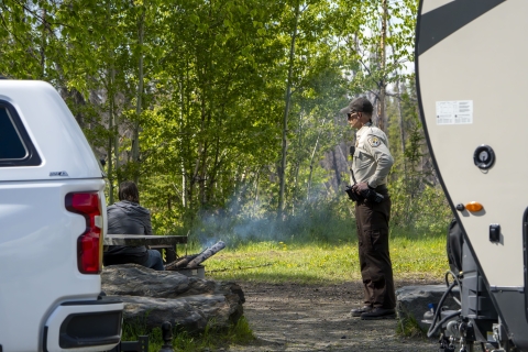 A uniformed Federal Wildlife Officer talks to a camper seated near a camp fire with trees in the background. They are framed into the image with a rear of a truck on the left and the front of a travel trailer on the right. 