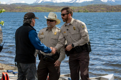 Two uniformed Federal Wildlife Officers meet the Secretary of the Interior Burgum in front of a lake with canoes on the shoreline and a large burn scar on the other side of the lake. Snow covered mountains cover the horizon. One of the officers is shaking hands with the Secretary. 