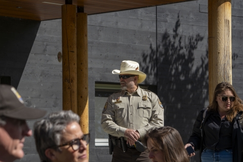 A uniformed Federal Wildlife Officer stands overwatch during a VIP visit. The visitors center of the Kenai National Wildlife Refuge covers the background. Out of focus in the foreground are three people taking part in the VIP visit. 