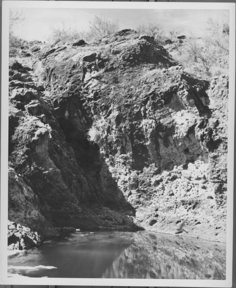 View of a pool of water, foreground, with a craggy, rocky natural wall behind it. The wall features a few crevasses and nooks and crannies.