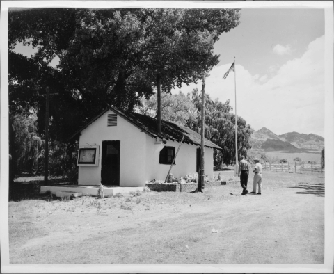 view of a small building with white stucco walls and a high-pitched roof. The end of the building facing the camera features a door and I framed box beside the door the has "BULLETINS" on the top frame. The side of the building features a window and a door. An exhaust pipe of some sort rises vertically from the left corner of the roof. To the right of the building is a tall metal flag pole. Two men, right center, are walking toward the building. A large tree towers over the building.