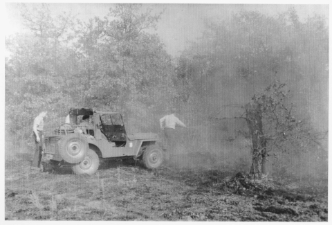 View of a jeep from the left side, and slightly behind; the vehicle is parked on the edge of a wooded area. A man (wearing a broadbrimmed hat) stands in front of the jeep, using what appears to be a hose to battle the fire in the trees and vegetation. Another man is sitting in the driver's seat of the jeep, while a third man stands at the back of the jeep. Smoke hangs in the air around the jeep. The tree in the right foreground appears to be burnt.