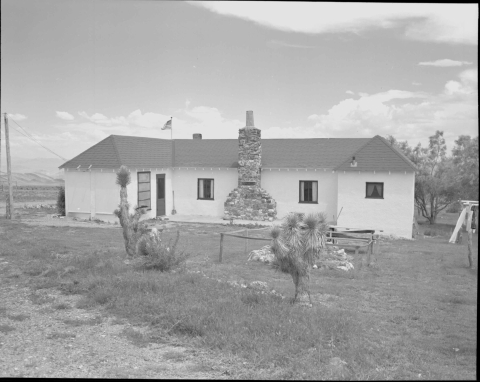 View of the single story residence/home that features while stucco walls and I high-pitched roof covered with darker shingles. Each end of the building are perpendicular to the long center section. A door is on the left end of the structure. In the center is a tall chimney made of rockwork, with two windows flanking it on either side. A small window is on the far right end. In the left foreground is gavel road. A couple of cacti are in the yard, along with a small area enclosed by a crude fence.