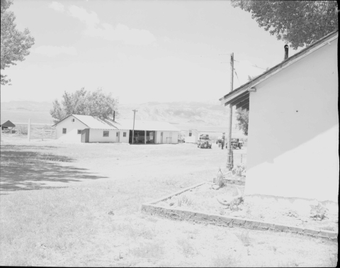 In the center background is a single-story building, with white walls, and a pitched roof with corrugated metal. In the center of the building appears to be a garage for motor vehicles (a truck is visible parked within it). The building also features about 5 windows, and two doors (on either side of the garage area). Another similar building is visible to the right of the previous described building. The corner of a third build (also of similar construction to the other two) is in the right foreground.