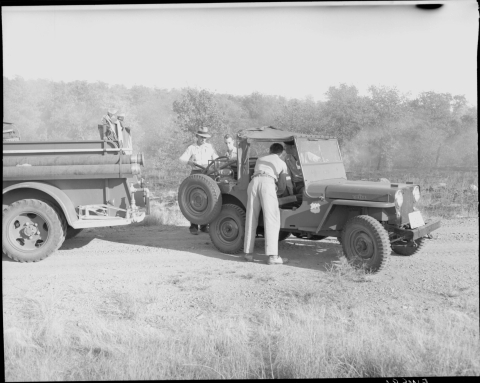 Three men gathered around a jeep, in the middle ground, with a fourth man sitting in the driver's seat, and looking to the back of the vehicle. One of the men is standing by the passenger's side of the jeep, while the remaining two stand at the back. On the left is the back end of a fire truck (the rest of the truck is out of frame). In the background are trees and brush. Wisps of smoke are visible against the vegetation.