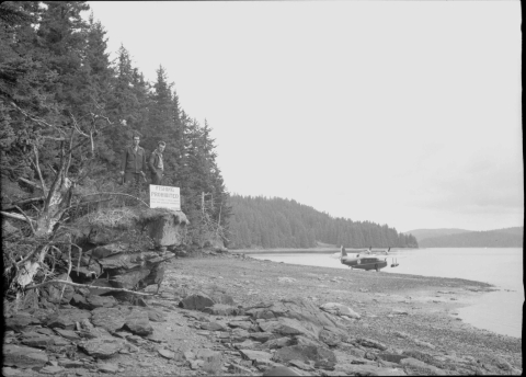 Two men standing on a short rocky ledge on the left, overlooking the shore of a bay (photo is taken from a position along the shore). In front of them is a sign that reads "FISHING PROHIBITED". In the middle distance parked along the shoreline is a large propeller-driven float plane. The shoreline features a narrow area that is clear of vegetation with a densely wooded area immediate beyond it. The shore stretches along the horizon; the opposite shore can also be seen in the right background.