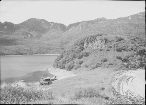 View from a high elevation, looking down on the shoreline of a lagoon in a bay. Visible along the shore is a small, crude structure, with a large, propeller-driven air plane. A mountain ridgeline rises behind the opposite shore of the lagoon.