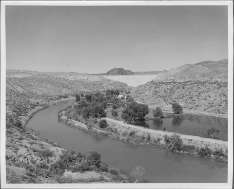 view taken from an elevated position, looking down on a river (the Rio Grande) as it curves from the center to the left and then to the right; in the distance is a large dam. At least two large fisheries ponds/pool are visible to the right of the river. Visible in the center distance (before the dam) is several hatchery buildings, nestled amongst a group of trees.