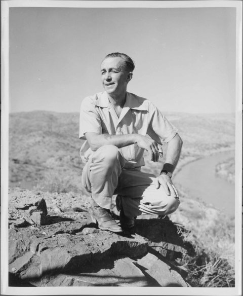 An older man, crouching down on an elevated area; visible behind him is valley with a watercourse running through it, on the right.