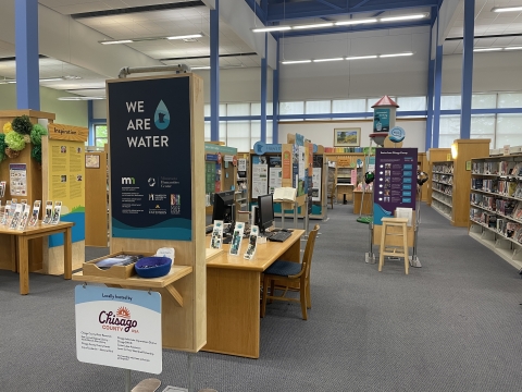Photo of We Are Water MN Exhibit as displayed at North Branch Area Library as part of hosting experience with Chisago County. In the foreground is a welcome panel with the We Are Water Logo, as well as logos for many state partners. The background shows many other exhibit pieces at a distance, including a 12 foot tall water tower that is part of the exhibit. 