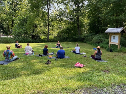 a group of people is seated on green grass with green trees and an information kiosk in the background