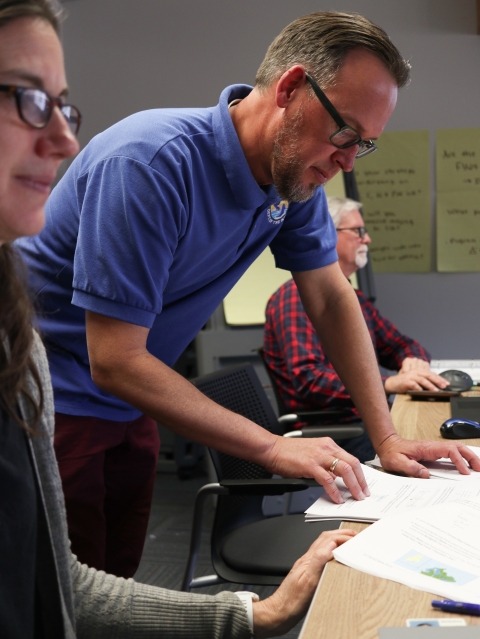 Man, standing. looking at a stack of papers, flanked by people working.