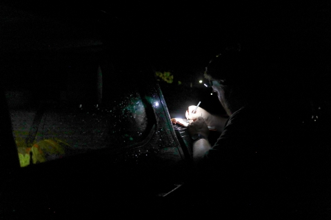 Man standing beside a truck, in the dark, using a headlamp while he writes in a notebook