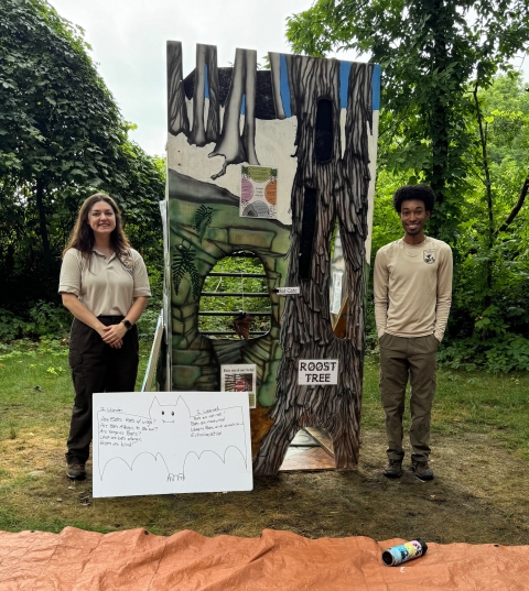 Two wildlife biologists in uniform standing next to a bat outreach display with a roost tree painted on it, and a whiteboard with a bat drawn on it 